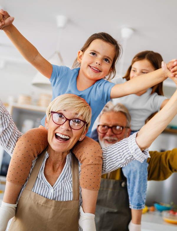 An endearing candid shot of an older couple, with two younger children sitting on their shoulders, all sharing joyful smiles and excitement during a playful moment.